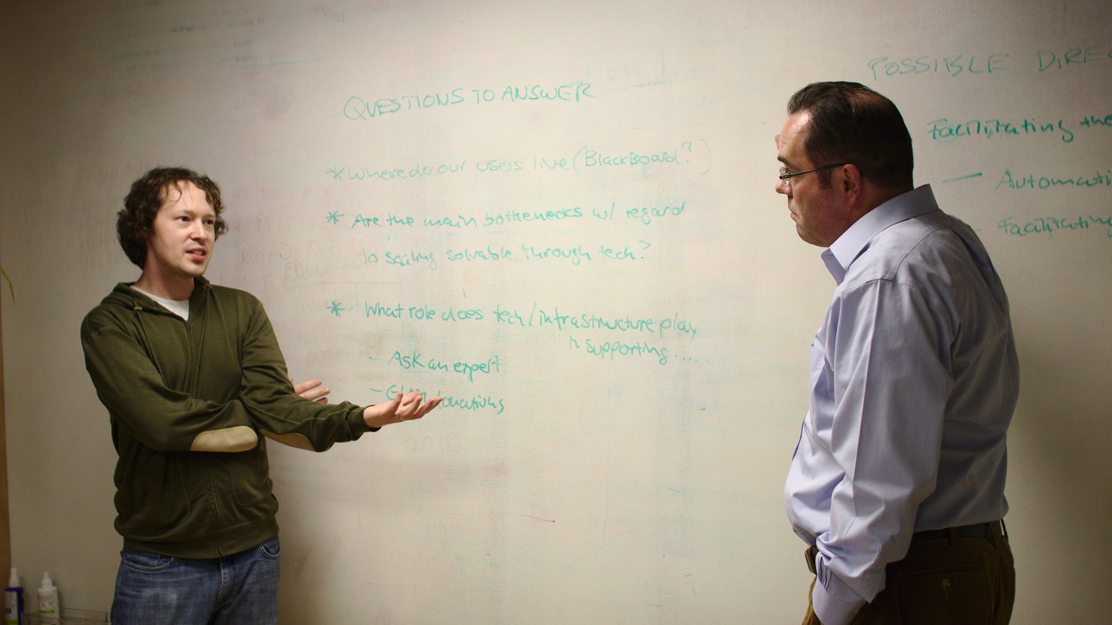 Two colleagues talking in front of a whiteboard covered in handwritten questions and action bullets