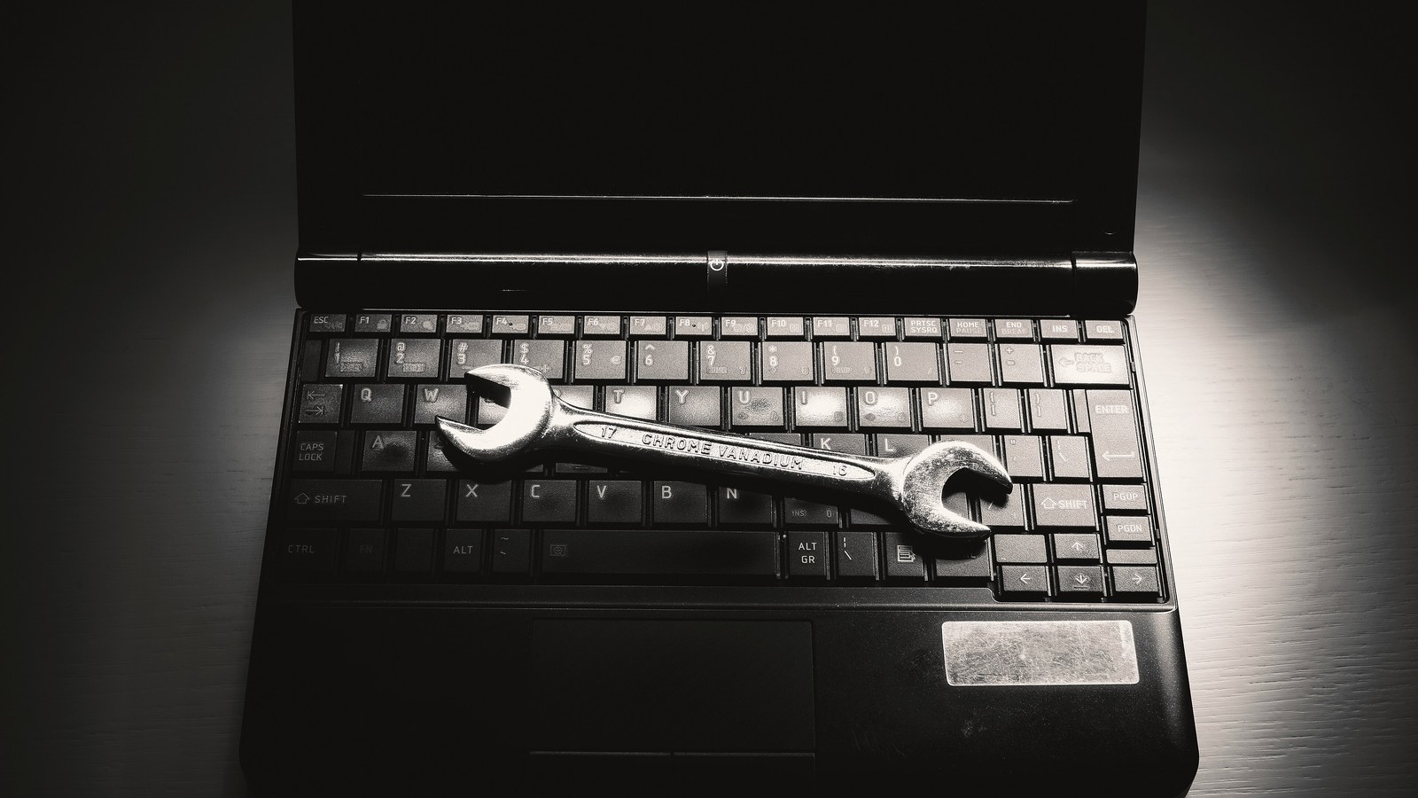 A metal wrench resting on the keyboard of an opened laptop, lit from one side against a dark background