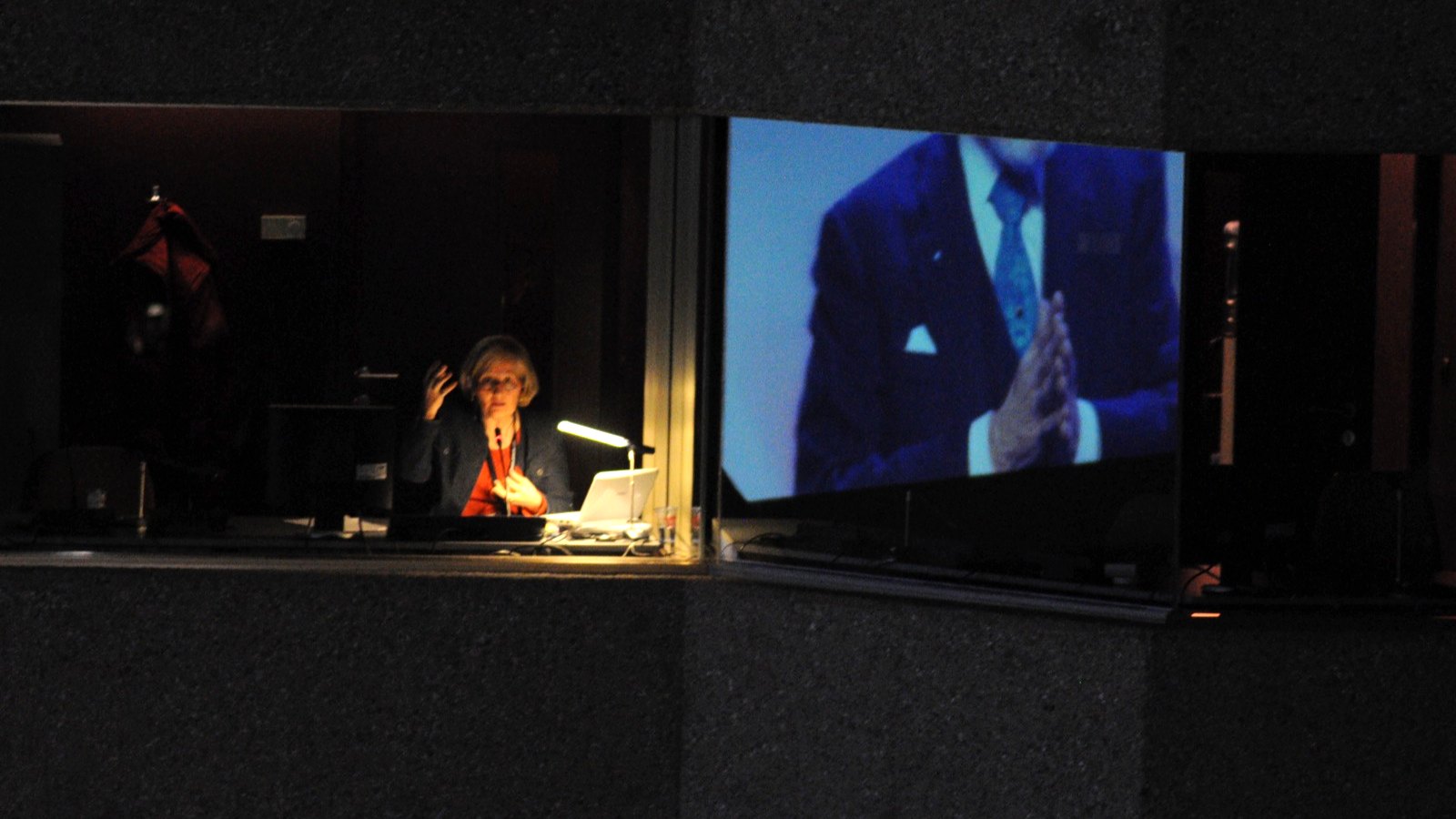 A simultaneous interpreter working in a lit booth at the back of a darkened conference hall, with the speaker she is interpreting shown on a large screen beside her